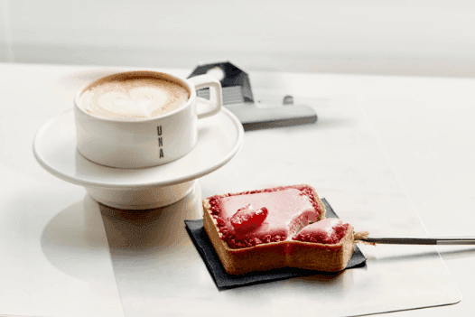 The photo shows a cup of coffee. Next to it lies a rectangular biscuit with raspberry glaze.